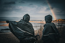 Load image into Gallery viewer, Two people in navy Carradice ponchos sitting on a beach with a rainbow in the background
